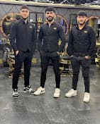 Three barbers in black uniforms posing together inside a modern barbershop with gold accents.