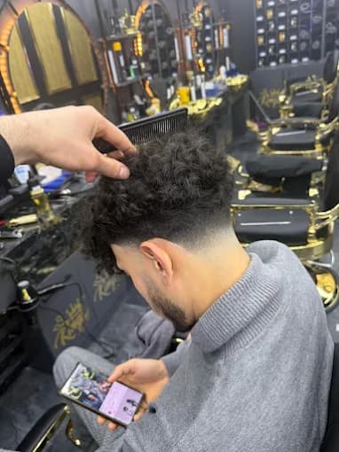 Barber combs a man's curly hair with a fresh fade in a stylish, gold-accented barbershop.